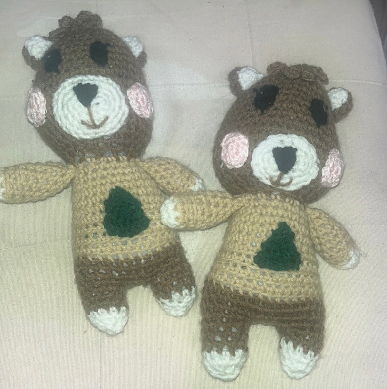 May include: Two handmade crochet bear dolls. The bears have brown heads with white and pink accents, black eyes, and a white muzzle. Their bodies are tan with a brown lower half and a green tree applique. The bears have white paws.