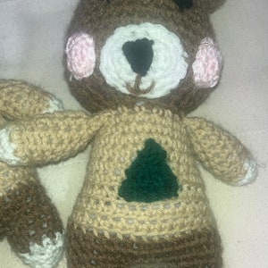 May include: Handmade crochet bear doll with a brown head, tan body, and dark brown legs. The bear has a white muzzle, pink cheeks, and a green tree design on its chest. The arms and feet have white accents.
