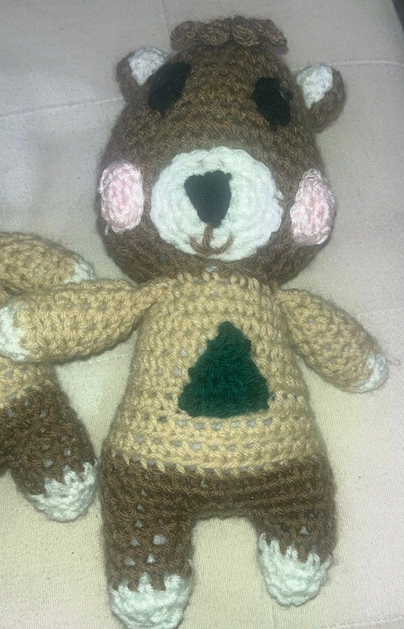 May include: Handmade crochet bear doll with a brown head, tan body, and dark brown legs. The bear has a white muzzle, pink cheeks, and a green tree design on its chest. The arms and feet have white accents.