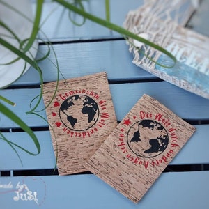 May include: Two cork coasters with a world map design and red text. The text on the left coaster reads "Gemeinsam die Welt entdecken" and the text on the right coaster reads "Die Welt durch andere Augen sehen".