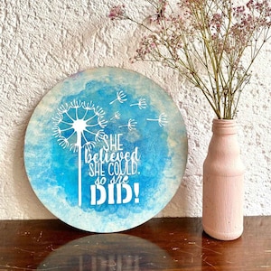 May include: A round wooden sign with a blue watercolor background and a white dandelion design. The sign has the text "She believed she could, so she did!" in white lettering.