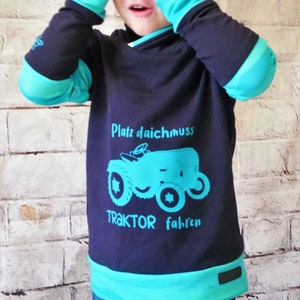 Könnte beinhalten: Ein dunkelblaues Langarmshirt mit türkisfarbenen Akzenten. Das Shirt zeigt ein Motiv eines blauen Traktors mit dem Text "Platz daichmuss TRAKTOR fahren".