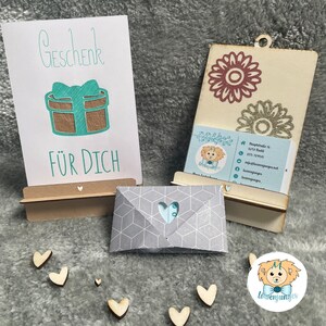 May include: A white card with the text "Geschenk für Dich" and a teal blue gift box with a teal blue bow. The card is on a wooden stand. There is a gray envelope with a white heart cutout. There are several small wooden hearts scattered around the image. There is a wooden sign with a business card that says "Löwenjunges".