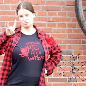 May include: A person wearing a black t-shirt with a red devil graphic and German text, paired with a red and black plaid shirt. The background features a brick wall and a wooden wagon wheel.