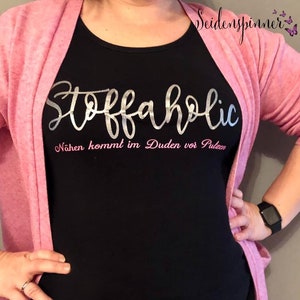 May include: A black t-shirt with silver glitter lettering that says "Stoffaholic" and pink lettering that says "Nähen kommt im Duden vor Pulzen".