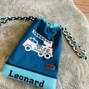 May include: A blue drawstring bag featuring a white fire engine graphic and the text "Brand Bekämpfer". The bag has a light blue base panel with the name "Leonard" embroidered in black. The drawstring is black and mint green.