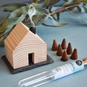 以下が含まれることがあります： 黒い煙突と煙が立ち上っている木製の家の香炉。香炉の隣には「ユーカリ」とラベル付けされた小さな香りのコーンのチューブがあります。薄い青色の表面にいくつかの茶色の香りのコーンがあります。