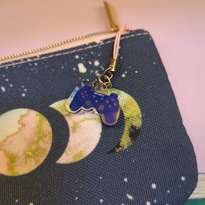 May include: A blue and clear acrylic charm in the shape of a video game controller, with the word "SONY" printed on it. The charm is attached to a gold-colored clasp and pink string. The charm is hanging from a navy blue cosmetic bag with a celestial design.