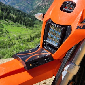 May include: Close-up of an orange motorcycle with a black and orange light bar mounted on the front. The light bar has the word "RIGID" printed on it. The motorcycle is parked in a mountainous area with green trees and grass in the background.