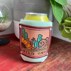 Könnte beinhalten: Ein hellblauer Leder-Getränkedosenkühler mit rosa und braunem Rand. Der Kühler zeigt ein handgemaltes Design mit einem Kaktus, einer Sonnenblume und einem Wirbel. Der Text "WILD ONE" ist auf dem Kühler geschrieben.