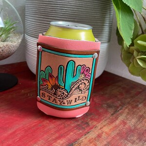 May include: A pink leather can cooler with a brown leather patch featuring a turquoise cactus, yellow flowers, and the text "STAY WILD".