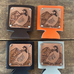 Könnte beinhalten: Vier Getränkedosenkühler in Braun, Orange, Schwarz und Grau. Jeder Kühler hat ein kupferfarbenes Quadrat mit einem detaillierten Reliefschnitzwerk eines Vogels und einer Berglandschaft, das mit silbernen Nieten befestigt ist. Die Kühler sind zum Kühlen von Getränken konzipiert.