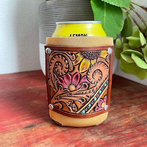 Puede incluir: Un enfriador de latas de cuero marrón con un diseño floral tallado que presenta girasoles, flores rosas y acentos turquesa. El enfriador tiene broches plateados y sostiene una lata amarilla con la palabra "LEMON" en ella.