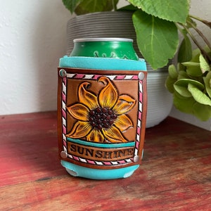 Könnte beinhalten: Ein türkisfarbener Leder-Getränkedosenkühler mit einem braunen Leder-Sonnenblumen-Design und dem Wort "SUNSHINE" in weißen Buchstaben. Der Kühler ist mit rosa und weißen Streifen verziert.