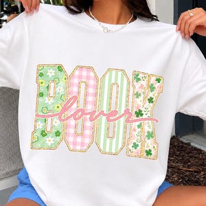 Könnte beinhalten: Ein weißes Kurzarm-T-Shirt mit dem Wort BOOK in großen Blockbuchstaben mit verschiedenen Mustern, darunter grüne Blumen, rosa Vichy-Karo, grüne Streifen und Kleeblätter, überlagert mit dem kursiven rosa Schriftzug lover.