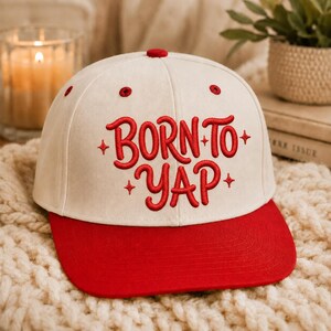 Peut inclure: Une casquette de baseball couleur crème avec une visière rouge et le texte brodé en rouge BORN TO YAP, agrémenté de petites étoiles à quatre branches. La casquette présente un bouton rouge sur le dessus et des œillets rouges.