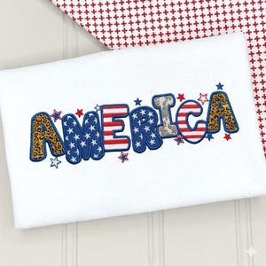 Könnte beinhalten: Eine weiße Stoffoberfläche mit dem gestickten Wort AMERICA in einem gemusterten Design. Die Buchstaben enthalten Leopardenmuster, Blau mit weißen Sternen sowie rote und weiße Streifen. Kleine gestickte Sterne umgeben den Schriftzug.
