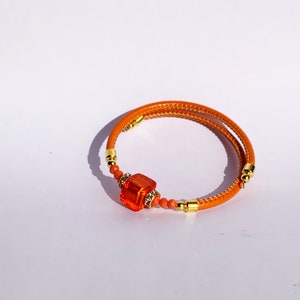May include: Orange spiral bracelet featuring a central orange square bead and smaller orange beads. The bracelet has gold-coloured accents. The bracelet is a fashion accessory, set against a white background.
