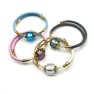May include: Four spiral bracelets in pastel colors: light blue, pink, cream, and gray. Each bracelet features a central square bead in a different color, accented with gold-toned metal and small clear beads. The bracelets are displayed on a white background.