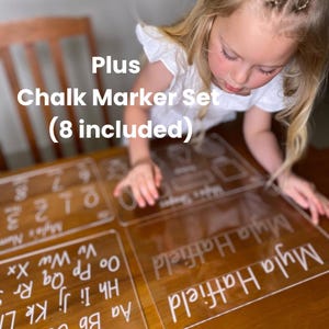 May include: Clear acrylic learning boards with white text, including the name "Myla Hatfield" and educational content. The image also shows the text "Plus Chalk Marker Set (8 included)". The boards are on a wooden surface.