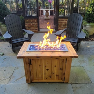 Può includere: Un tavolo da fuoco in legno con un vano di stoccaggio integrato. Il focolare è acceso con vetro fuoco blu ed è circondato da due sedie nere. Il focolare è di fronte a una serra.