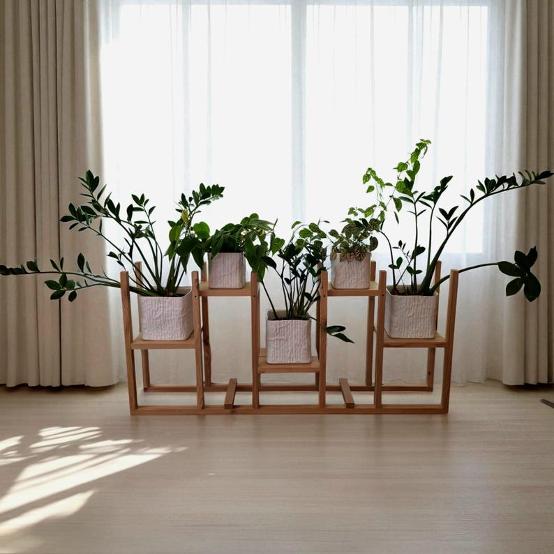 May include: A wooden plant stand with five square white planters, each holding a different variety of green plants. The stand is positioned in front of a window with sheer white curtains, creating a bright and airy atmosphere.