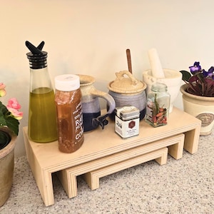 May include: A set of three unfinished wooden risers, each a different size, arranged in a tiered display. Various kitchen items are displayed on the risers, including a glass bottle, a honey bottle, and a small tin of seasoning.
