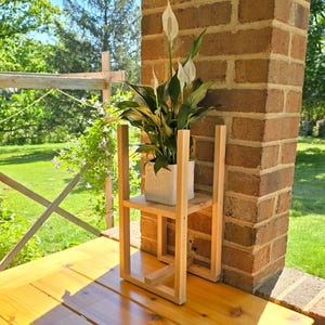 May include: A wooden plant stand with two shelves, holding a white square pot with a green plant and white flowers. The stand is made of light-colored wood and is placed next to a brick pillar. The background shows a wooden deck and greenery.