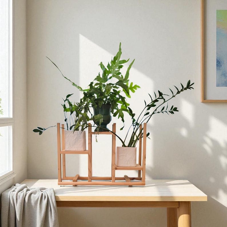 May include: A wooden plant stand with three sections, holding a dark green vase and two light pink square planters, each filled with green plants. The stand is on a light-colored wooden table, with a window and artwork in the background.