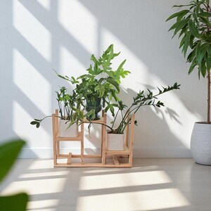 May include: A wooden plant stand with three levels, holding various potted green plants. The stand has a geometric design and is made of light-colored wood. The plants are in white and green pots, creating a natural, indoor aesthetic.
