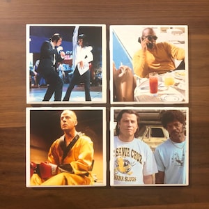 May include: Four square ceramic coasters featuring images from popular movies. The first coaster shows a man in a black suit and a woman in a white shirt dancing. The second coaster shows a man in a yellow shirt talking on a phone. The third coaster shows a man in a yellow jumpsuit holding boxing gloves. The fourth coaster shows two men, one in a blue shirt with the text "Santa Cruz" and the other in a white shirt with a cartoon image.