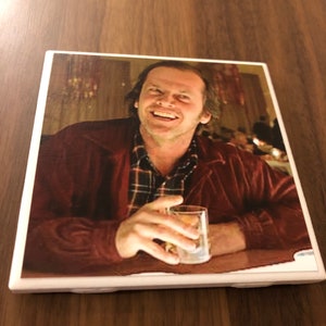 May include: A ceramic coaster featuring a close-up image of Jack Nicholson as Jack Torrance from the movie "The Shining". He is smiling and holding a glass of liquid.