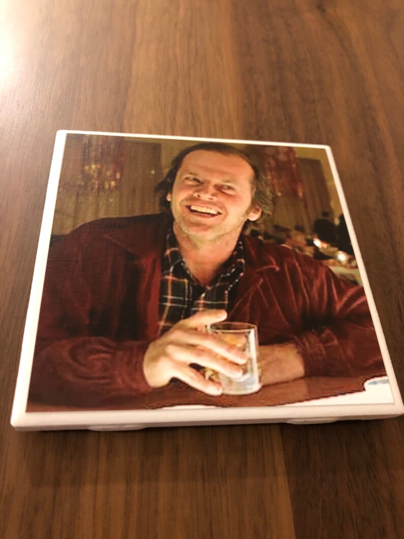 May include: A ceramic coaster featuring a close-up image of Jack Nicholson as Jack Torrance from the movie "The Shining". He is smiling and holding a glass of liquid.