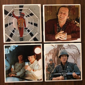 May include: Set of four ceramic coasters featuring iconic movie scenes. The first coaster shows a person in a spacesuit walking down a hallway. The second coaster shows a man with a glass of liquid in his hand. The third coaster shows two men sitting with glasses of milk. The fourth coaster shows a man in a military uniform holding a rifle.