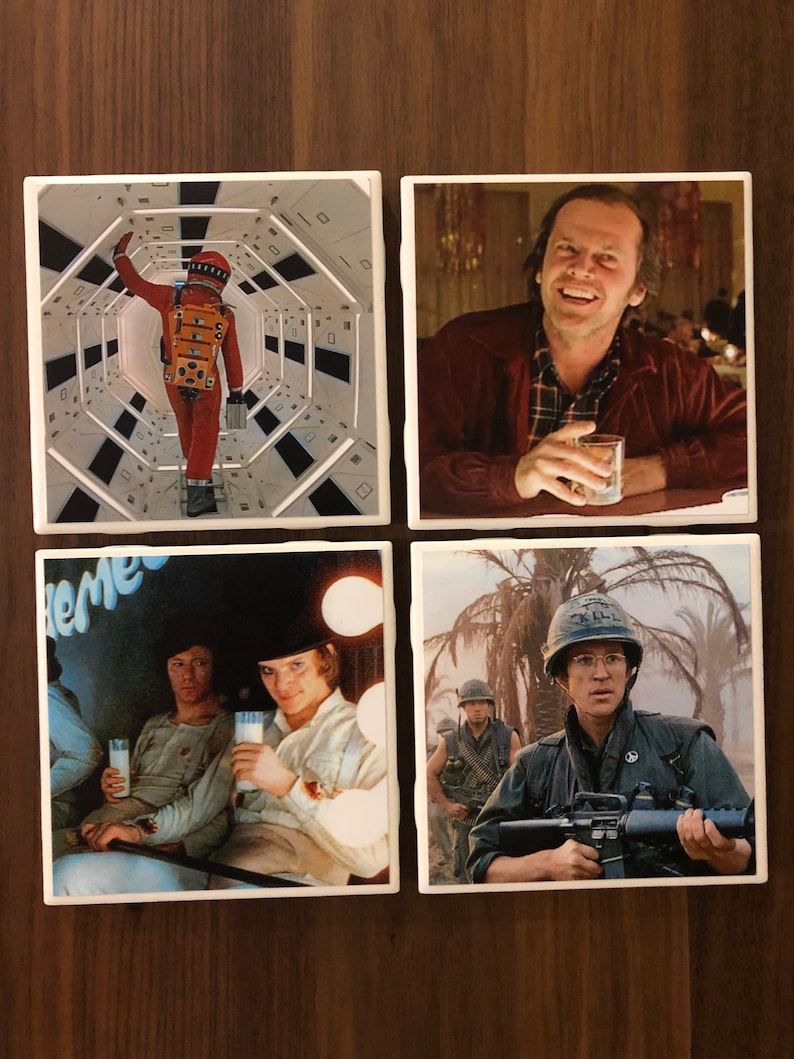 May include: Set of four ceramic coasters featuring iconic movie scenes. The first coaster shows a person in a spacesuit walking down a hallway. The second coaster shows a man with a glass of liquid in his hand. The third coaster shows two men sitting with glasses of milk. The fourth coaster shows a man in a military uniform holding a rifle.