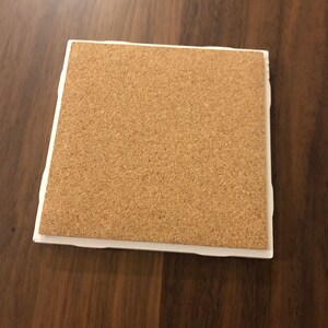 May include: A square cork coaster with a white border. The coaster is made of natural cork and has a slightly rough texture.