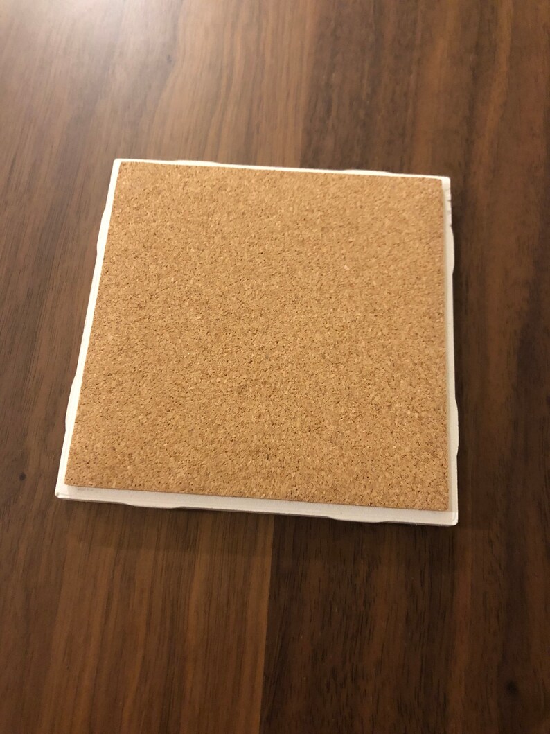May include: A square cork coaster with a white border. The coaster is made of natural cork and has a slightly rough texture.