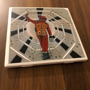 May include: A white square coaster with a raised border and a printed image of an astronaut in an orange spacesuit walking down a long, white corridor.