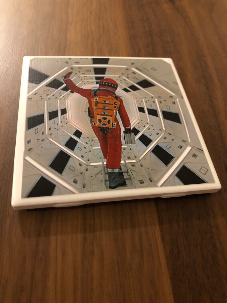 May include: A white square coaster with a raised border and a printed image of an astronaut in an orange spacesuit walking down a long, white corridor.