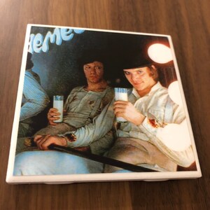 May include: A vintage style coaster featuring a black and white image of two people drinking milk. The image is set in a dark room with a blue and white sign that reads "The" in the top left corner.