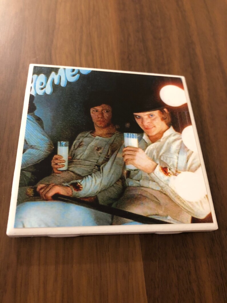 May include: A vintage style coaster featuring a black and white image of two people drinking milk. The image is set in a dark room with a blue and white sign that reads "The" in the top left corner.
