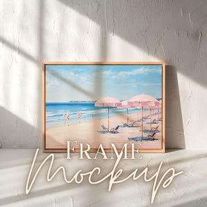 Op de afbeelding: Een ingelijst schilderij van een strandscène met roze parasols en strandstoelen. Het schilderij is in een licht houten lijst en hangt aan een witte muur.