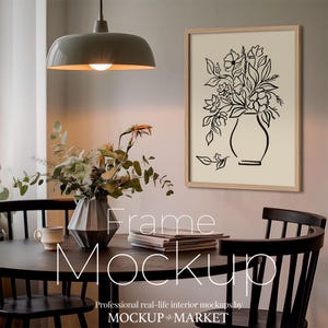 May include: A framed black and white line drawing of flowers in a vase, hanging on a wall above a wooden table with two chairs. The table has a vase of flowers and a stack of books on it. The text "Frame Mockup" is written on the table.