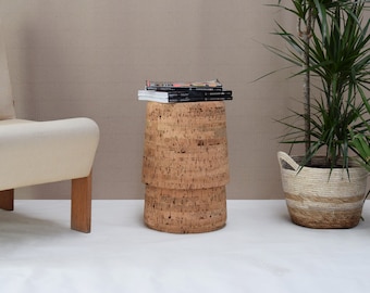 May include: A cylindrical side table made of cork, with a stack of books on top. The table is light brown and has a textured surface. A beige armchair and potted plants are in the background.