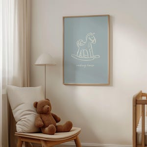 May include: A framed art print featuring a white line drawing of a rocking horse on a light blue background. The words "rocking horse" are written below the image. The print is displayed in a light-colored wooden frame. A brown teddy bear sits on a wooden chair.
