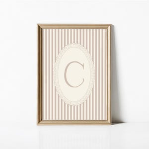 Heirloom-inspired monogram nursery wall art featuring the initial 'C' within a lace frame against classic pink and white stripes.  A natural fit for interiors inspired by the feminine, refined aesthetic of spaces thoughtfully curated by Anthropologie and Studio McGee.
