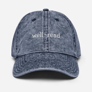 May include: A faded blue denim baseball cap with the embroidered text "well-read" in white. The cap has a curved peak and an adjustable strap. The text is centrally positioned on the front of the cap.