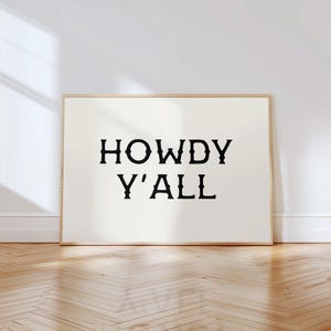 May include: A framed art print with the words "HOWDY Y'ALL" in bold, black letters. The print has a cream-colored background and a light brown wooden frame. The artwork is displayed on a white wall above a wooden floor.