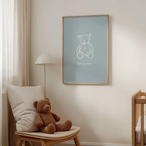 May include: A framed poster with a light blue background features a white line drawing of a teddy bear and the words "teddy bear" below it. A brown teddy bear sits on a wooden chair with a cream-colored pillow. A floor lamp and a crib are also visible.