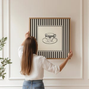 May include: A framed wall art print featuring a black and white illustration of a teacup and saucer. The artwork is set against a cream background and framed with a black and white striped border. The frame is being held up by a person.